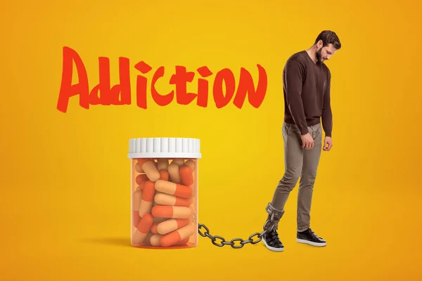 Giovane depresso in piedi e guardando in basso, incatenato a enorme vaso di plastica di pillole su sfondo ambrato con grande titolo rosso Addiction . — Foto Stock