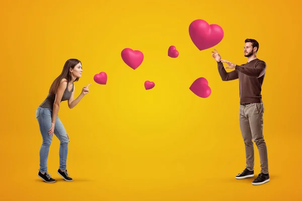 Vue latérale du jeune couple sur fond ambré jouant avec de mignons cœurs de Saint-Valentin qui flottent dans l'air, l'homme essayant d'attraper un cœur . — Photo