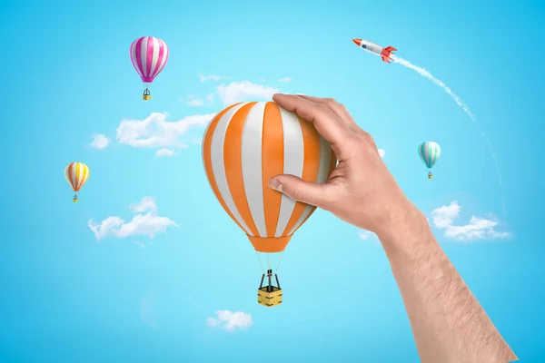Mão segurando balão de ar quente branco laranja com foguete de prata e balões de ar quente no fundo do céu azul — Fotografia de Stock