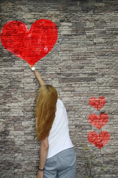 Una chica joven pinta un corazón rojo acuarela en la pared de piedra, th —  Fotos de Stock