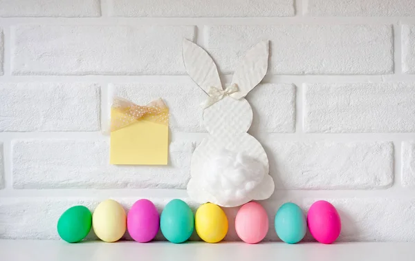 Easter white bunny with colored eggs with sticky note against white brick wall background.