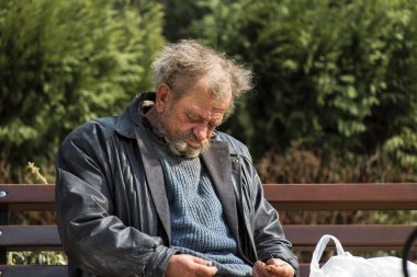 Zolochiv, Ukraine - April 10, 2018: Homeless man sitting asleep on a bench in the city center along Markian Shashkevich Street, near the Church of the Resurrection of the Lord clipart