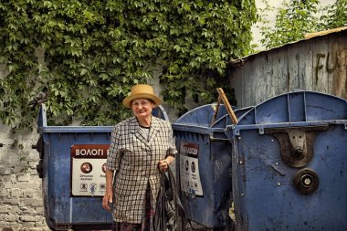 Vinnitsa, Ukraine - May 9, 2018:  Elderly woman hoarder in a straw hat and checkered suit rummaging in a dumpster clipart