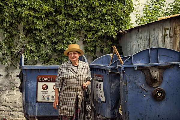 Vinnitsa, Ukraine - May 9, 2018:  Elderly woman hoarder in a straw hat and checkered suit rummaging in a dumpster