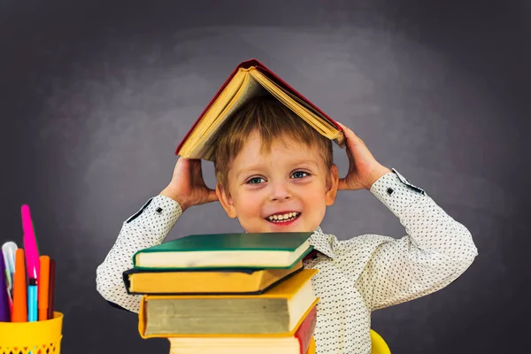Przedszkolak siedzi przy biurku na tle tablicy i trzyma książkę na głowie.. — Zdjęcie stockowe