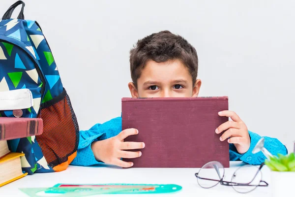 Um menino de cabelos escuros com uma camisa azul em uma mesa com livros . Fotos De Bancos De Imagens