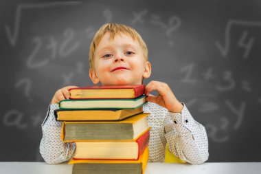 Sarışın bir çocuk kitaplarla dolu bir masada oturuyor. Anaokulu çocuğu oturuyor.