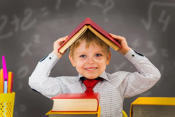 未就学児の少年は黒板の後ろに机に座って頭に本を抱えています — ストック写真