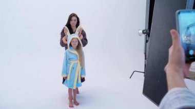 Elinde fotoğraf makinesi olan kadının, gri arka planda bornozlu neşeli anne ve kızının fotoğraflarını çekişi.