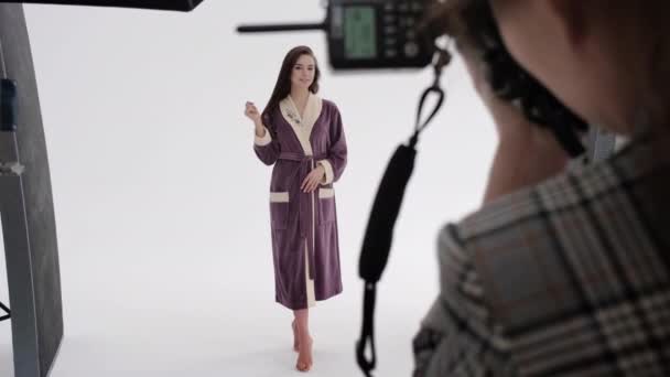 Back view of woman using photo camera to shoot female model in pink bathrobe during photoshoot in professional studio — Stock Video