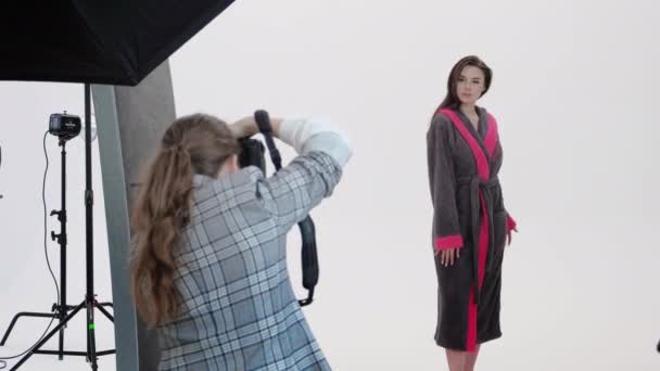 Vista posterior de la mujer utilizando la cámara de fotos para disparar modelo femenino en albornoz rosa durante la sesión de fotos en un estudio profesional — Vídeos de Stock
