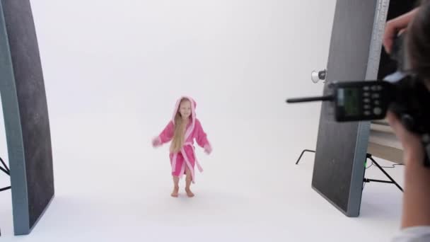 Nierozpoznawalna osoba z kamerą robiąca zdjęcia podekscytowanej dziewczynie w szlafroku skaczącej na białym tle w studio — Wideo stockowe