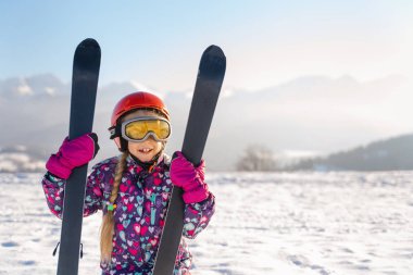 Little girl wearing white helmet and outwear holding mountain skis in sunlight