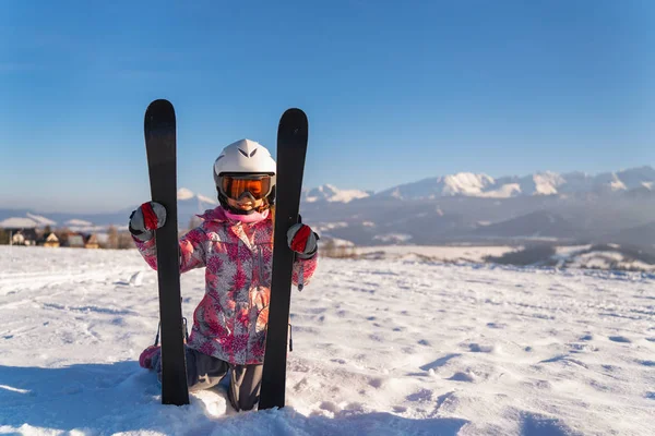 Little girl wearing white helmet and outwear holding mountain skis in sunlight