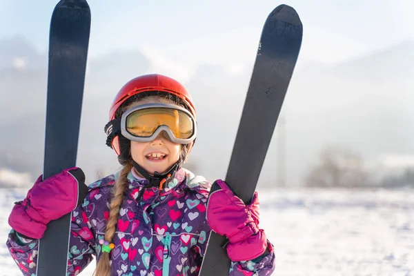 Little girl wearing white helmet and outwear holding mountain skis in sunlight