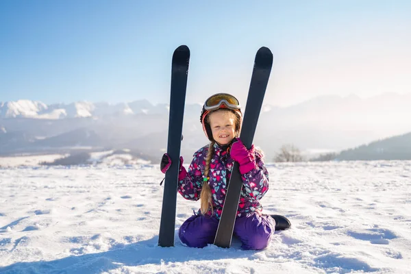 Little girl wearing white helmet and outwear holding mountain skis in sunlight