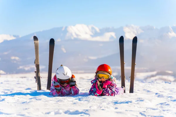 Kinderen in helmen en brillen liggend op besneeuwde terrein met ski's in de buurt op de achtergrond van de bergen Stockfoto