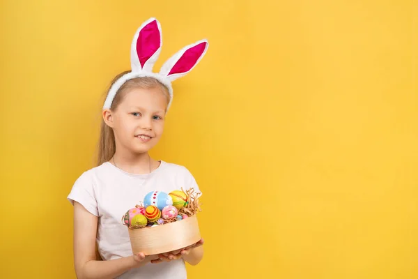 Amazed little girl with bunny ears looking at camera and showing painted egg during Easter celebration against yellow background — Stock Photo, Image