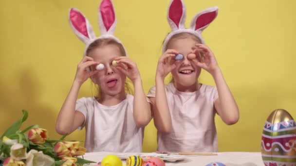 Hermanas lindas con orejas de conejo manteniendo huevos de codorniz coloridos cerca de los ojos y mostrando lenguas mientras se prepara para la celebración de Pascua contra el fondo amarillo — Vídeos de Stock