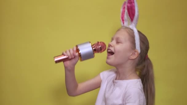Очаровательная маленькая девочка с кроличьими ушами и микрофоном, улыбающаяся и смотрящая в камеру, поющая песни во время празднования Пасхи на желтом фоне — стоковое видео