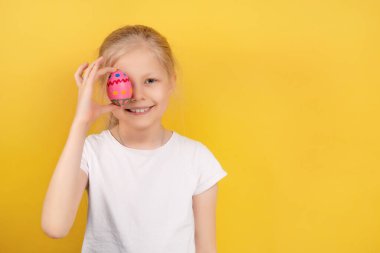Tavşan kulaklı tatlı bir kız. Renkli yumurtayı göz önünde tutuyor ve sarı arka planda Paskalya kutlaması sırasında kameraya gülümsüyor.