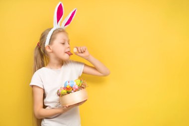 Tavşan kulaklı hayret verici küçük kız kameraya bakıyor ve sarı arka planda Paskalya kutlaması sırasında boyalı yumurta gösteriyor.