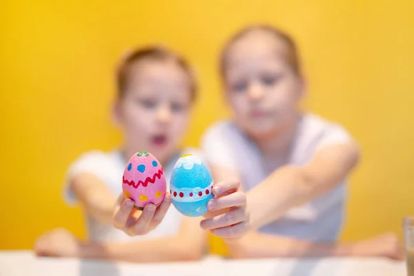 Irmãzinhas bonitos com ovos coloridos sentados à mesa com símbolos paleta e Páscoa contra fundo amarelo — Fotografia de Stock
