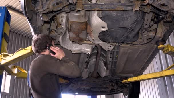 Mechanic guy with phone repairing rusty exhaust system. — Stock Video