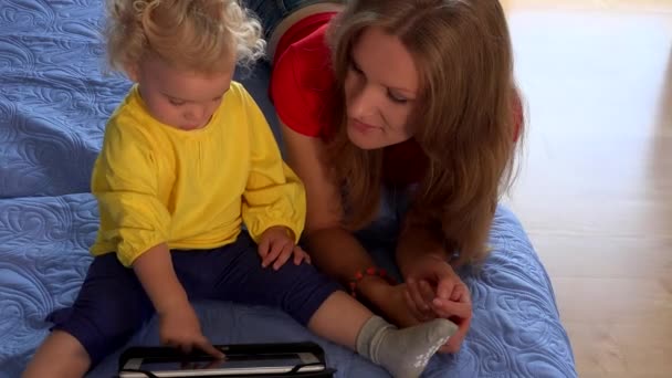 Bebé rubia con mujer morena madre usando la tableta en la cama azul — Vídeo de stock