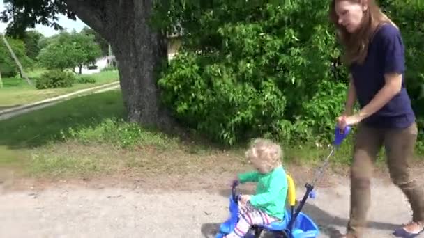 Fiatal nő édesanyja szőke kisgyermek lány nyomja a kis tricikli. — Stock videók