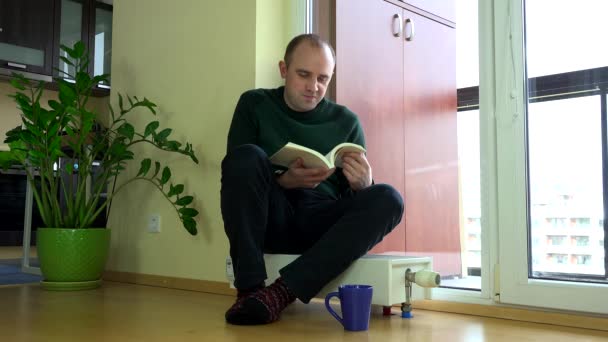 Mladý muž číst knihu a teplý na chladič doma. — Stock video