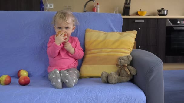 Hermoso niño niño pequeño sentado en el sofá y comiendo fruta de manzana grande . — Vídeos de Stock