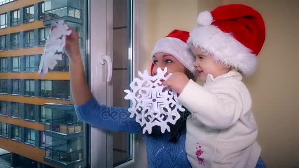Kärleksfull kvinna och barn flicka med rött santa hattar lim papper snöflingor på fönster — Stockvideo