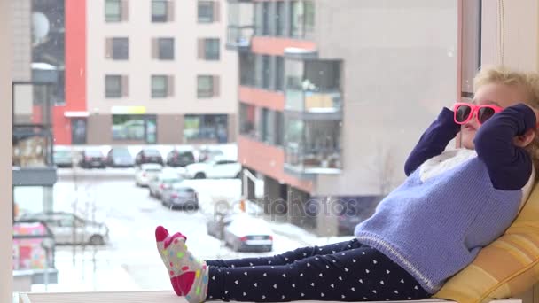 Menina bonito com óculos de sol rosa sentar na soleira da janela e flocos de neve cair fora — Vídeo de Stock