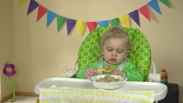 Chica divertida comer gachas con cuchara y las manos sentadas en la silla de alimentación del bebé . — Vídeos de Stock