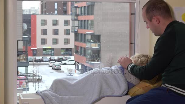 Cuidado padre acaricia a su hijo acostado en el radiador cerca de la ventana. Nieve cayendo — Vídeo de stock