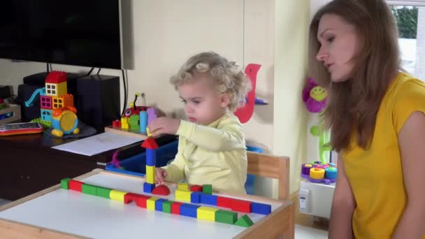 Niña con madre jugando ladrillos de madera de colores cerca de una pequeña mesa en casa — Vídeo de stock