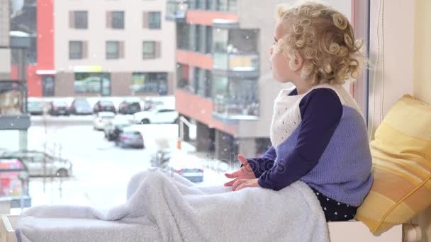 Niño curioso mirando caer nieve por la ventana en invierno — Vídeos de Stock