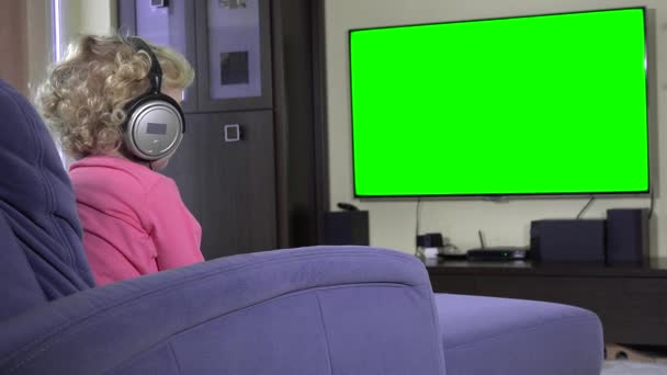 Liten unge tittar på TV med stora hörlurar. Gröna chroma key skärmen — Stockvideo