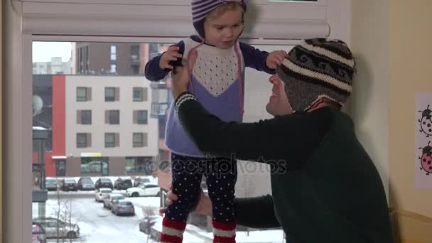 Отец с сыном в теплых шляпах веселятся у окна. Семья готовится выйти на улицу — стоковое видео
