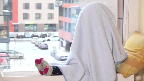 Verspieltes Mädchen mit Handtuch im Haar, das auf einem Heizkörper am Fenster sitzt. Schneesturm — Stockvideo