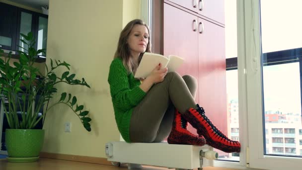 Jolie femme assise sur le chauffage de la maison et lire le livre en hiver . — Video