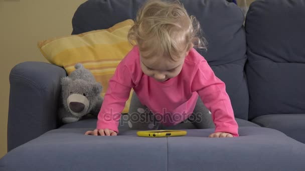 Dulce niña caucásica jugando con el teléfono inteligente solo en el sofá azul . — Vídeos de Stock