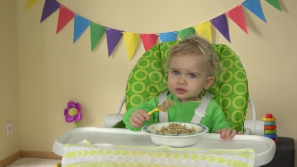 Hungrig baby flicka med sked sitter vid utfodring stol och äter gröt från plattan — Stockvideo