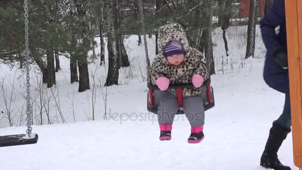 Nő anya és a baba lány swing a téli játszótér. A hó hófúvás. 4k — Stock videók