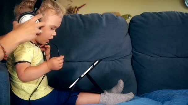 As mãos de mulher põem grandes fones de ouvido na filha com a cabeça de computador de pastilha . — Vídeo de Stock