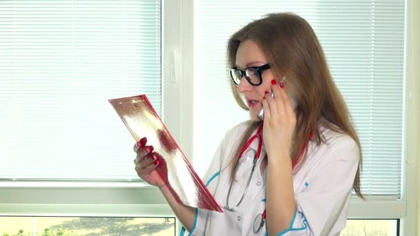 Jovem médica bonita com pasta de arquivo falando no telefone celular com seu paciente — Vídeo de Stock