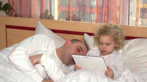 Солодка дівчинка-малюк за допомогою планшетного комп'ютера прокидається сплячий тато чоловік в ліжку — стокове відео