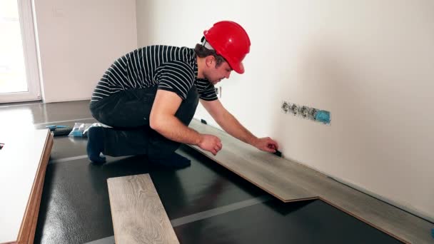 Рабочий с красным шлемом, устанавливающий деревянный пол в новом доме — стоковое видео