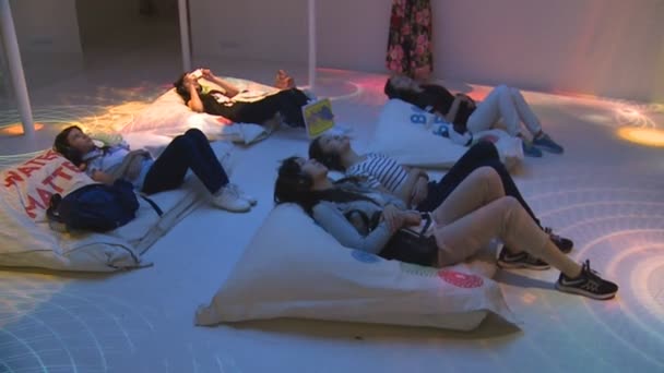 Visitantes personas con auriculares se encuentran en enormes almohadas en el suelo y ver la pantalla de televisión — Vídeo de stock